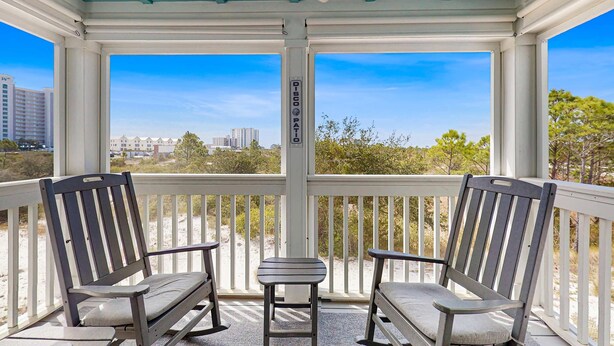 A covered outdoor seating area with two rocking chairs and a small table, overlooking a beach and buildings. - Gorgeous townhouse, plenty of space, lots of games, shared pool, close to beach (Perdido Key)