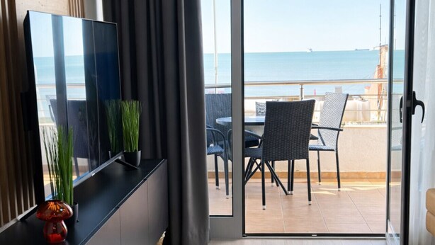 Een moderne kamer met een balkon met uitzicht op zee, voorzien van een eettafel gedekt met borden en glazen, een televisie op een zwarte standaard, en een potplant.
