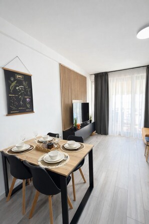 Un comedor moderno con una mesa de madera dispuesta para cuatro, sillas negras y un mueble de televisión con marco negro al fondo.