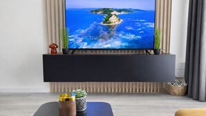 Een moderne woonkamer met een flatscreen televisie bevestigd aan een zwarte entertainmentkast, een klein bijzettafeltje met decoratieve objecten, en uitzicht op een kustlandschap.