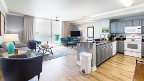 Ein modernes Wohnzimmer mit einer Couch, einem Couchtisch und einem Fernseher. Es gibt eine Küche mit Mikrowelle, Herd und Schränken. Der Raum verfügt über Holzböden und ein großes Fenster mit Vorhängen.