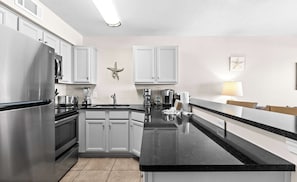 A modern kitchen with stainless steel appliances, white cabinetry, a central island, and a starfish decoration on the wall. - Oceanfront Condo w/ AC, WiFi, Shared Pool, & Seasonal Beach Service Rentals (Destin)