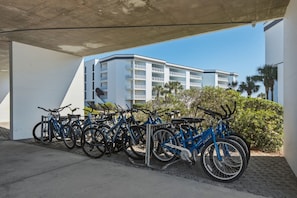 A bike parking area with multiple bicycles under a concrete overhang, a white building with balconies in the background, and some greenery. - Attractive home with spacious balcony, pool view & pickleball - near beach (Santa Rosa Beach)