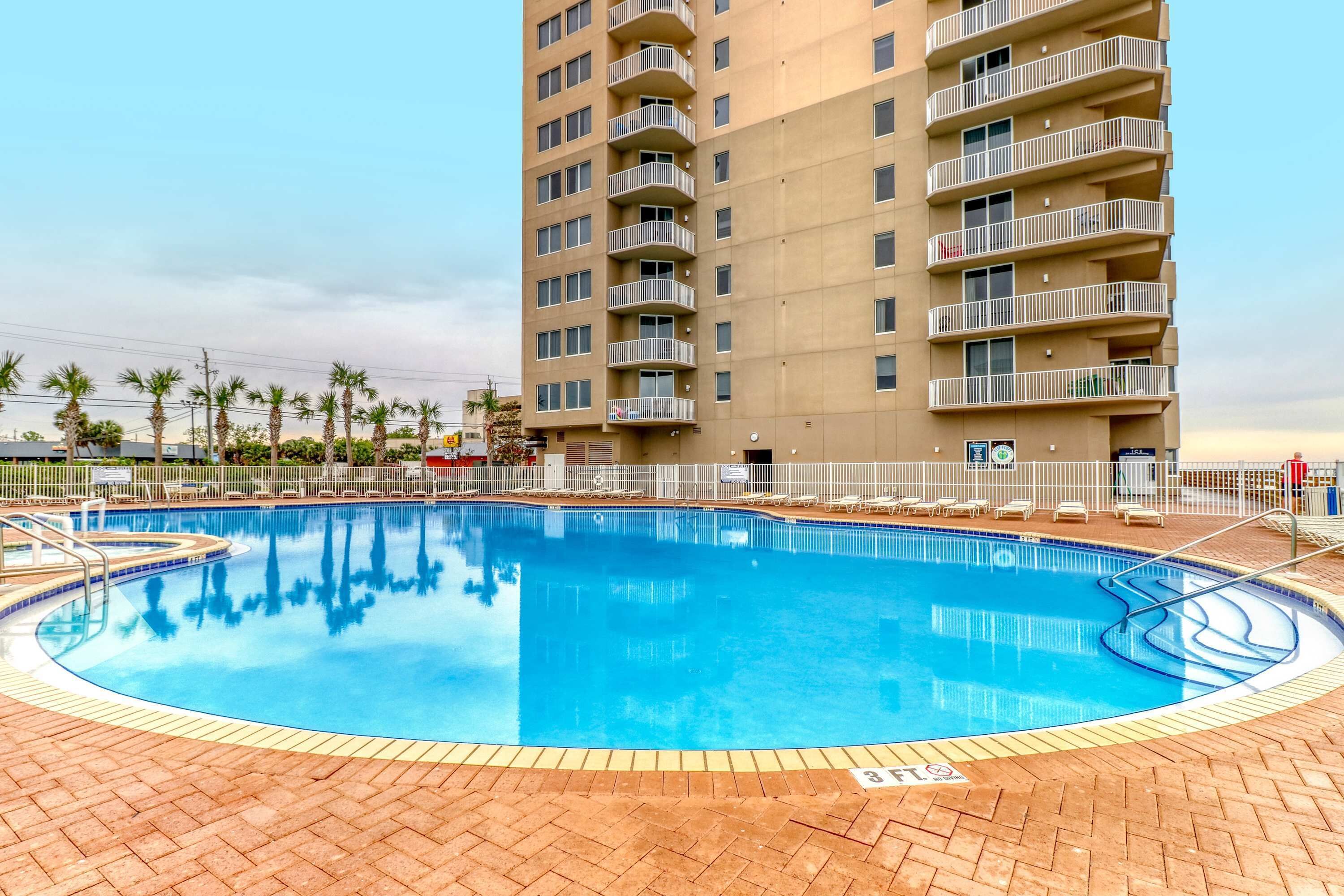 Une piscine bordée de briques, entourée de chaises longues et située devant un immeuble imposant doté de balcons.