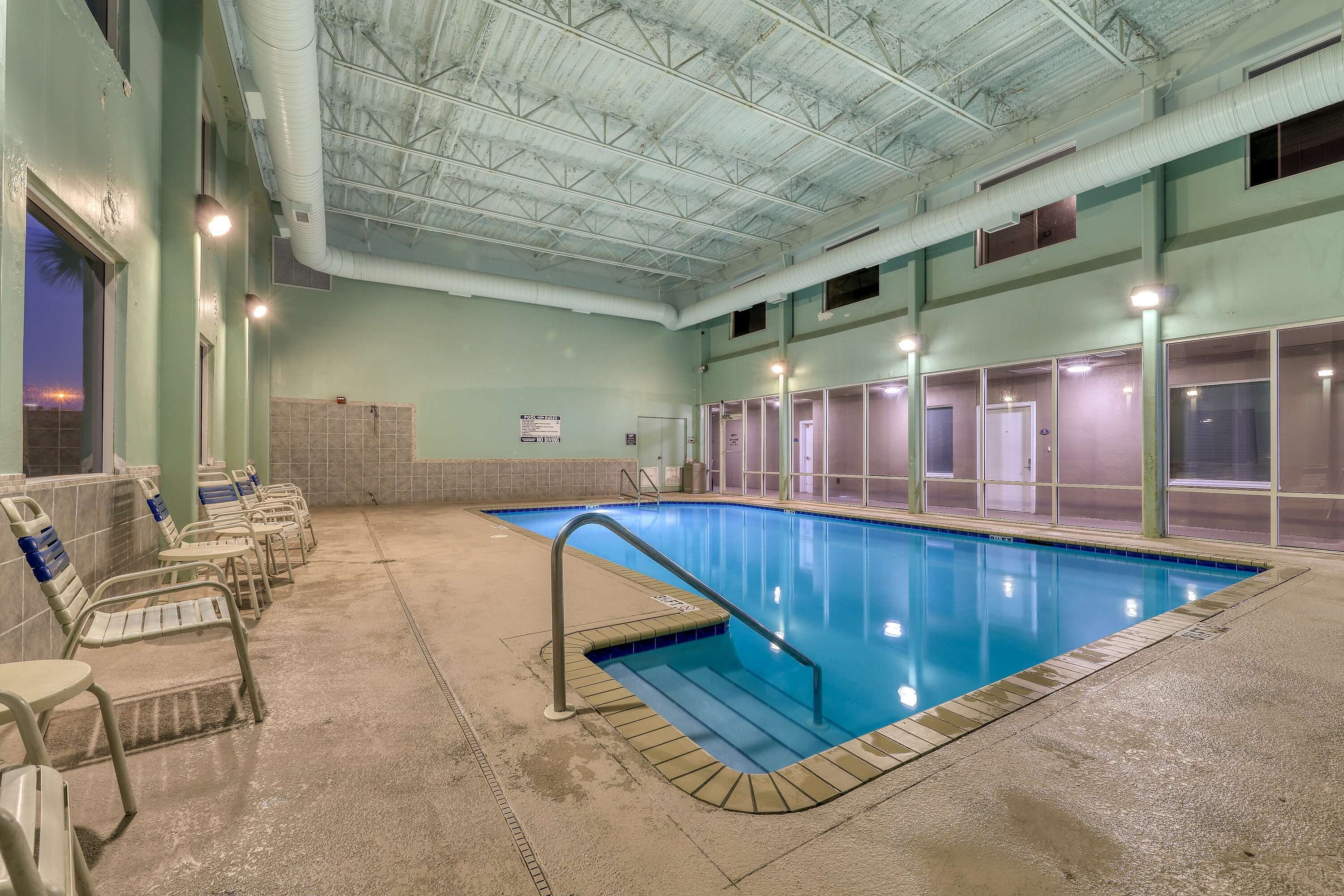 An indoor swimming pool with a clear water surface, surrounded by a concrete floor and a poolside area with chairs.