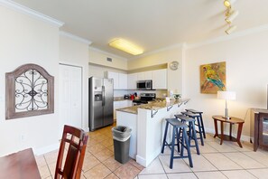 A modern kitchen with white cabinets, a stainless steel refrigerator, a microwave, and a granite countertop. There are two bar stools and a small table with a lamp. - Gulf-front getaway with water views, pools, hot tub, fast WiFi, & central AC (Panama City Beach)
