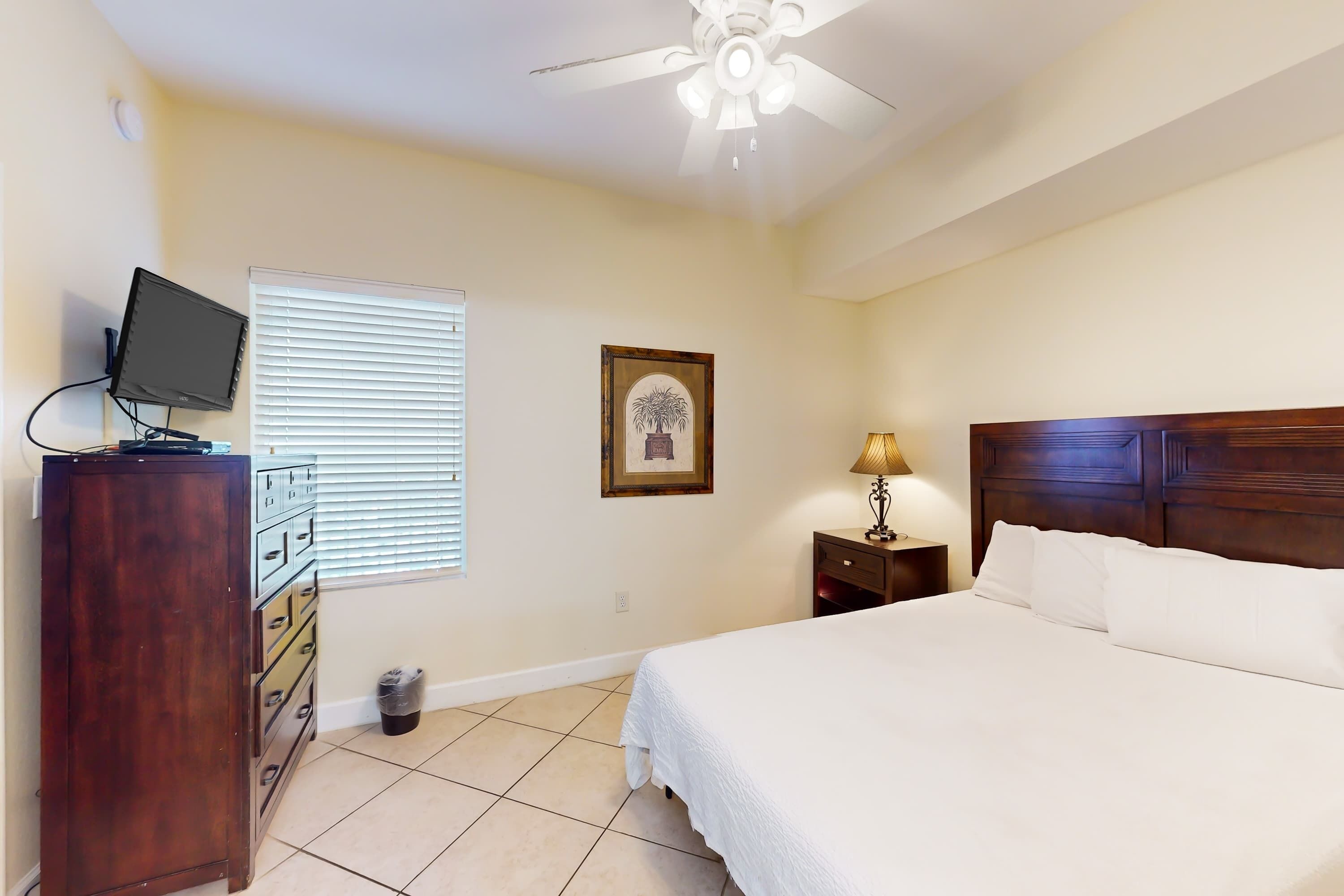 A bedroom with a bed, a dresser, a ceiling fan, a lamp, and a window with blinds.