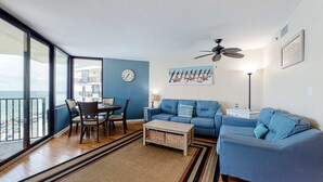 Ein Wohnzimmer mit blauem Sofa, einem Holztisch, einem Esstisch mit Stühlen, einem großen Fenster mit Blick auf den Strand und einem Deckenventilator.