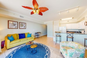 A modern living room with a yellow sofa, a patterned armchair, and a blue rug. A kitchen with white cabinets and a bar area is visible in the background. - Picture-perfect Gulf front condo with gym, pools, hot tub & sauna (Panama City Beach)