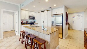 Una cucina moderna con isola centrale, sgabelli da bar, mobili bianchi, elettrodomestici in acciaio inossidabile e pavimento piastrellato.