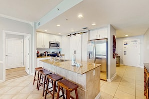 A modern kitchen with a central island, bar stools, white cabinets, stainless steel appliances, and a tiled floor. - Seasonal Beach Chairs included in Gulf Front Condo rental! (Panama City Beach)