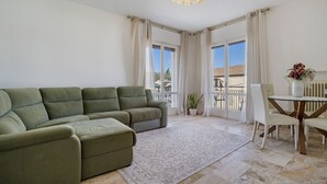 En stue med en modulsofa, et spisebord med stole, en lysekrone og store vinduer med gardiner.