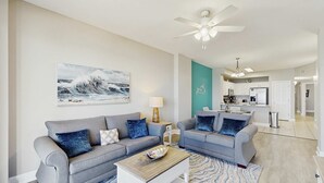 A living room with two grey sofas, a coffee table, and a painting of waves on the wall. - Water-view beachfront condo with pool - steps to the beach (Panama City Beach)