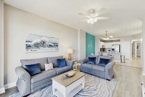 A living room with two grey sofas, a coffee table, and a painting of waves on the wall. - Water-view beachfront condo with pool - steps to the beach (Panama City Beach)