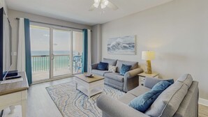 A spacious living room with a large window, a ceiling fan, a sofa, a coffee table, and a painting on the wall. - Water-view beachfront condo with pool - steps to the beach (Panama City Beach)
