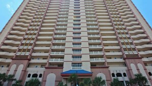 A high-rise building with multiple balconies, a central entrance, and a blue roof. - Water-view beachfront condo with pool - steps to the beach (Panama City Beach)