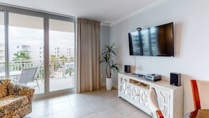 Ein modernes Wohnzimmer mit großem Fernseher, einem weißen Schrank mit Lautsprechern, einer Couch, einem Topf mit Pflanze und Blick auf einen Balkon mit einem Stuhl.