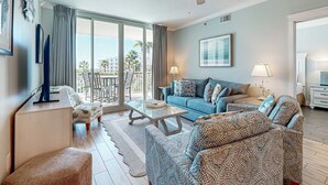 A modern living room with a sofa, a coffee table, and a TV. There is a balcony with a view of palm trees and buildings. - Stunning, Spacious Condo At Waterscape Steps To Beach Lazy River (Fort Walton Beach)
