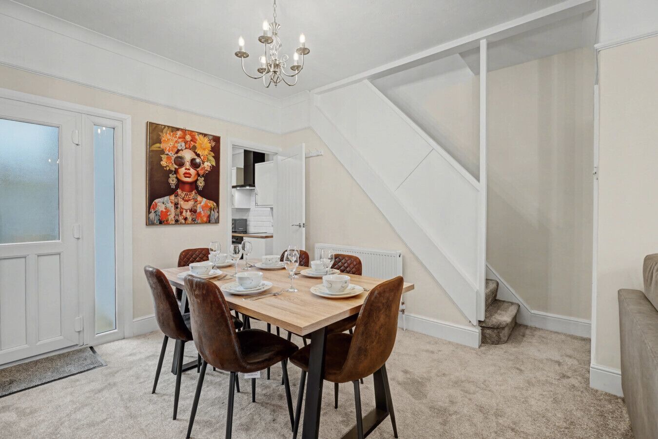 A dining area with a wooden table set for four, surrounded by brown chairs. A chandelier hangs from the ceiling, and a staircase is visible to the right.