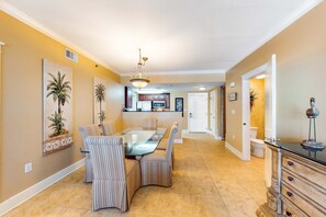 A dining area with a glass table and striped chairs, a kitchen in the background, and a hallway leading to a bathroom. - Cute, coastal condo with lazy river, Gulf view, hot tub & gym - near the beach (Fort Walton Beach)