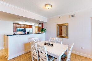 Ein Essbereich mit weißem Tisch und Stühlen, eine Küche im Hintergrund und ein Spiegel an der Wand.