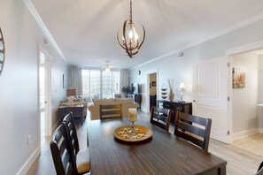 A dining area with a wooden table, chairs, and a chandelier. There is a sofa and a television in the background. - 3rd Floor Welcoming, Coastal Condo At Waterscape Playground, 2 Hot Tubs (Fort Walton Beach)