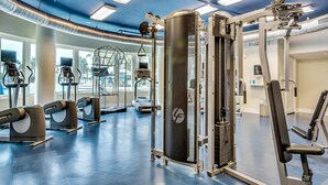 Ett gym med olika typer av träningsutrustning, inklusive löpband och styrketräningsmaskiner, och stora fönster som släpper in naturligt ljus.