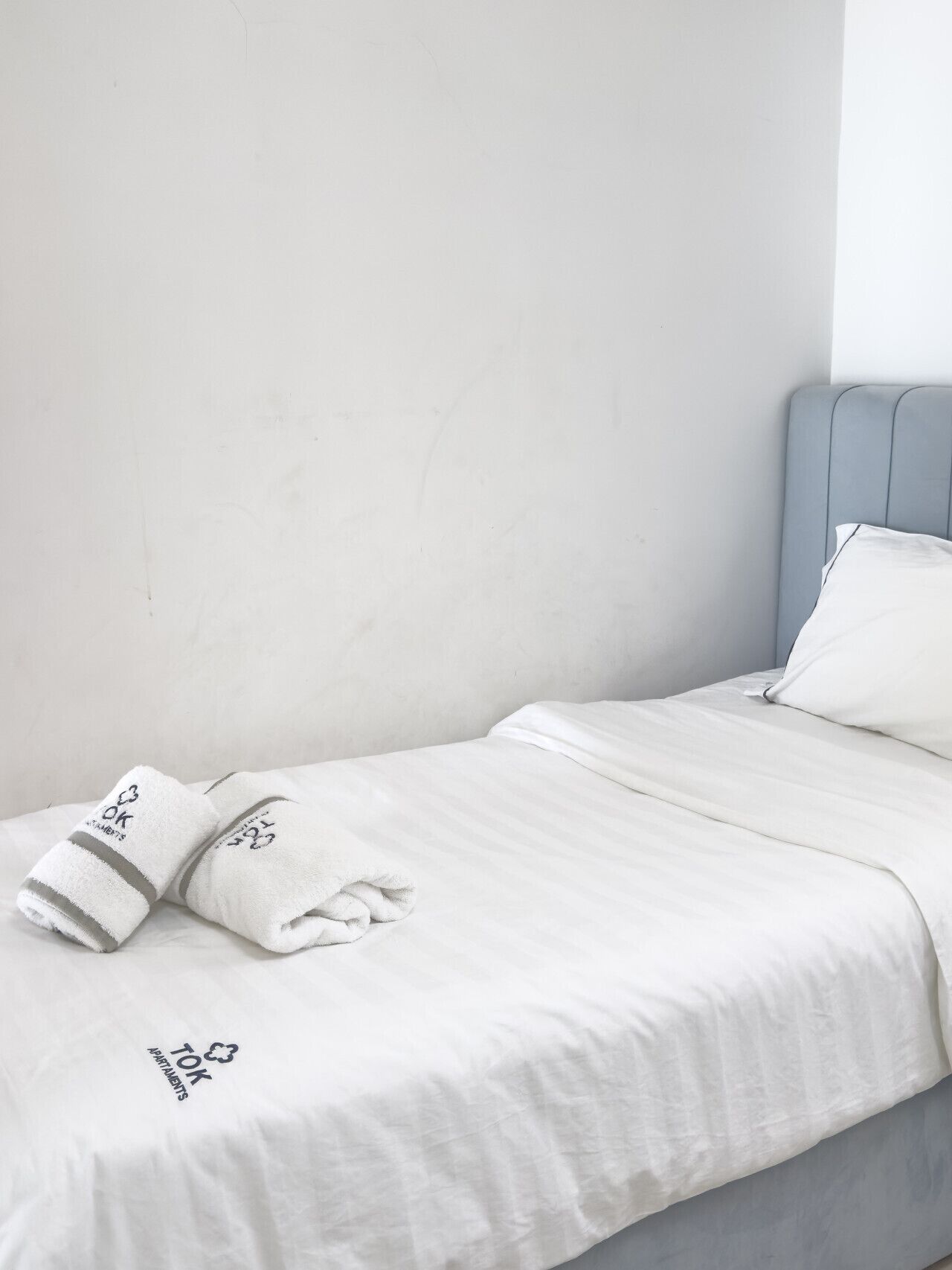 A single bed with a white bedspread and two folded towels with a logo.