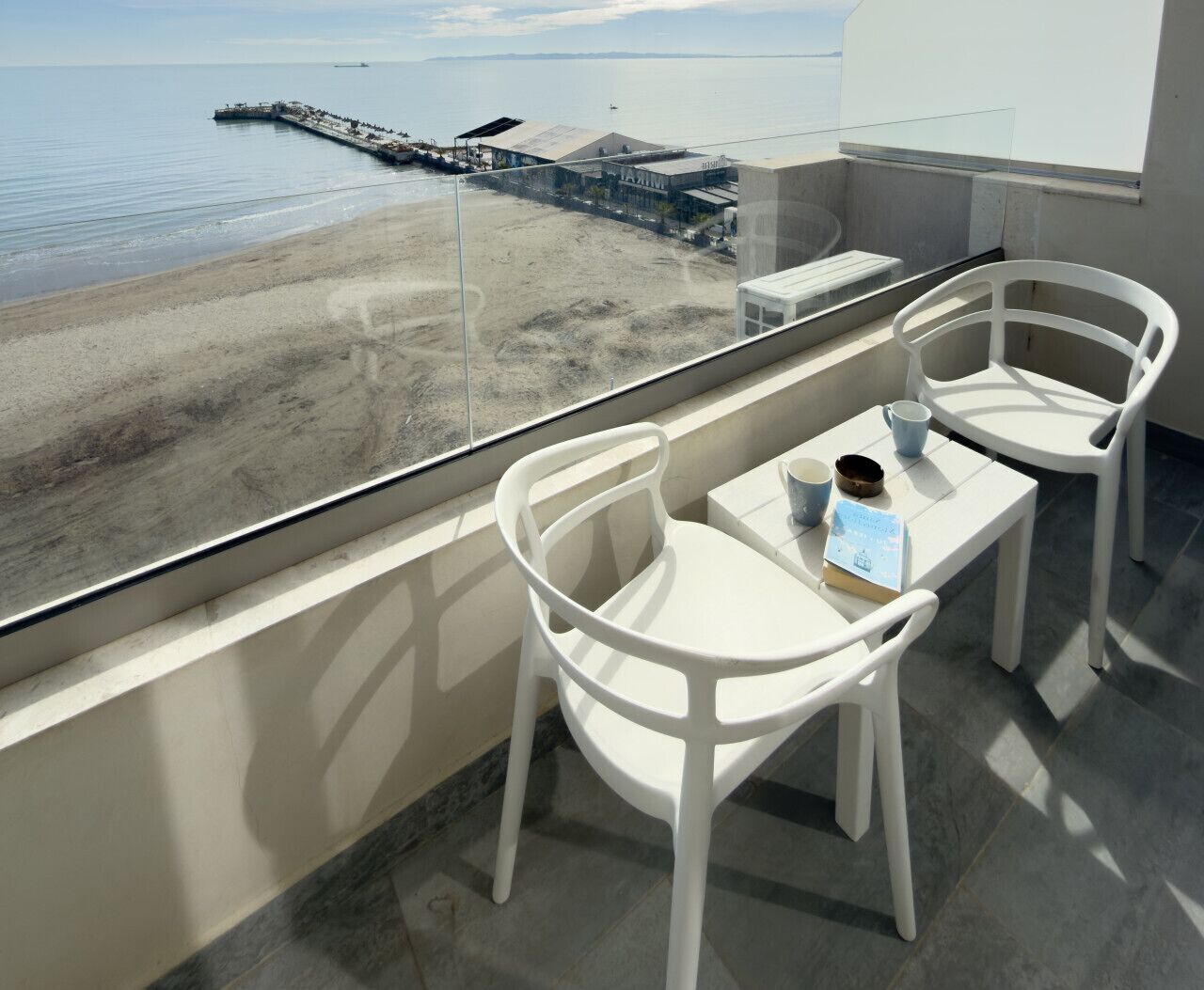 Ein Balkon mit zwei weißen Stühlen und einem Tisch, der Blick auf einen Strand und einen Steg freigibt.