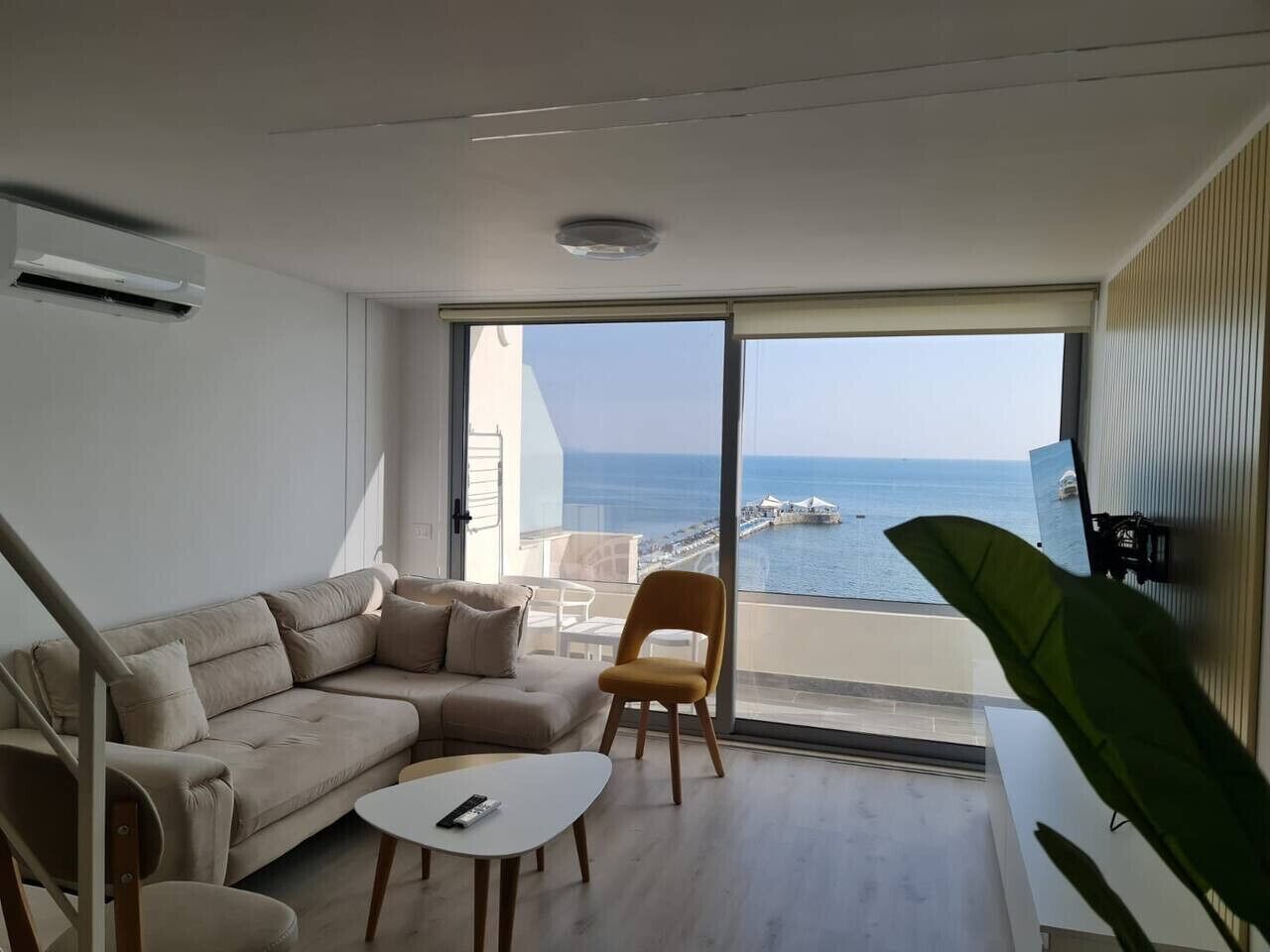 A modern living room with a beige sofa, a small white table, and a yellow chair. A large window offers a view of the sea and a distant cityscape.