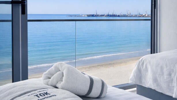 A hotel room with a bed, a window with a view of the sea and a port, and a towel on the bed.
