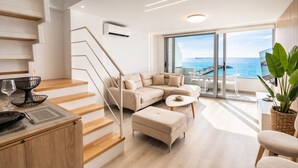 A modern living room with a beige sofa, a coffee table, and a staircase leading to an upper level with a view of the sea.