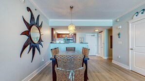 A dining area with a wooden table and chairs, a chandelier, and a sun-shaped mirror on the wall. - Beach-Chic Condo On Okaloosa Island Waterfall, Lazy River (Fort Walton Beach)