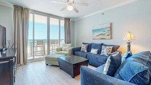 A spacious living room with a large window, a ceiling fan, a sofa set, and a coffee table. - Beach-Chic Condo On Okaloosa Island Waterfall, Lazy River (Fort Walton Beach)
