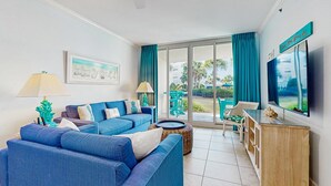 A living room with a blue sofa, a TV, and a balcony with blue curtains. - Bright, beachy condo At Waterscape w children's playground, 2 hot tubs (Fort Walton Beach)