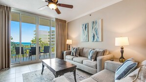 A living room with a sofa, coffee table, and two lamps. There is a ceiling fan and a view of the outside through large windows. - 3rd Floor Picturesque Condo On Okaloosa Island Waterfall, Lazy River (Fort Walton Beach)