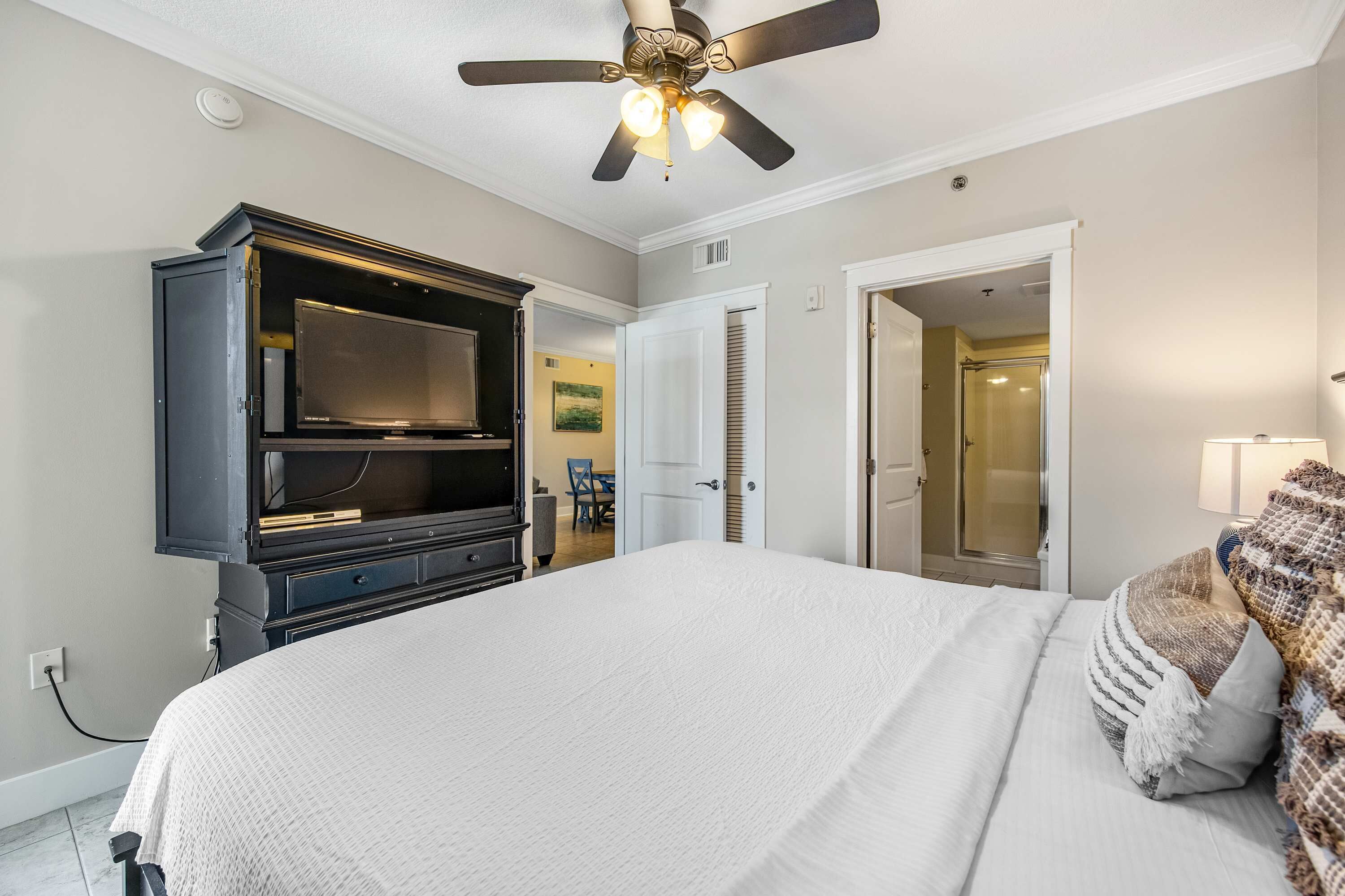 A bedroom with a large bed, a dark wooden dresser, a ceiling fan, and a door leading to a bathroom.