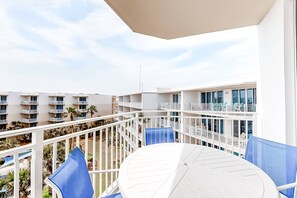 Varanda com mesa branca e cadeiras azuis, com vista para uma piscina e outros edifícios do hotel.