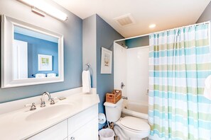 Casa de banho com lavatório branco, espelho e sanita. Existe uma cortina de chuveiro com padrão riscado.
