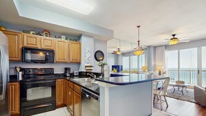 A modern kitchen with wooden cabinets, a built-in microwave, and a dishwasher. There is a dining area with a table and chairs, and a view of the outdoors through large windows. - Elegant & spacious condo with incredible Gulf-front views, two pools & gym (Panama City Beach)
