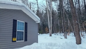 Una casa de un piso con fachada gris y contraventanas negras, rodeada por un paisaje nevado y árboles.