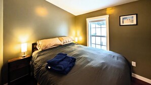 Un dormitorio con cama, mesita de noche, ventana y un cuadro en la pared.
