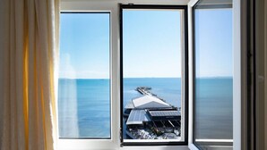 A room with a large window overlooking a coastal area, featuring a building with solar panels and a pier. - Sea view -Blue Wave Two (Durrës)