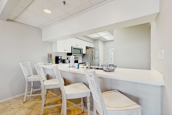 A kitchen with white cabinets, a microwave, a coffee maker, and a sink. There are two white chairs with cushioned seats. - Airy beachfront condo with balcony views, lagoon pool, tennis & pickleball (Seacrest)
