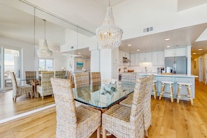 A modern kitchen with white cabinets, a stainless steel refrigerator, and a glass dining table with wicker chairs. - Lavish High Pointe condo with resort pool, amazing ocean view & pickleball (Seacrest)