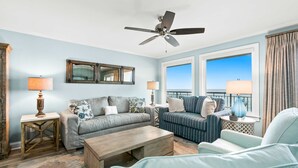 A living room with a ceiling fan, two sofas, a coffee table, and a view of the ocean through large windows. - Oceanfront condo with pool, ocean view, washer&dryer, & high-speed WiFi (Fort Walton Beach)
