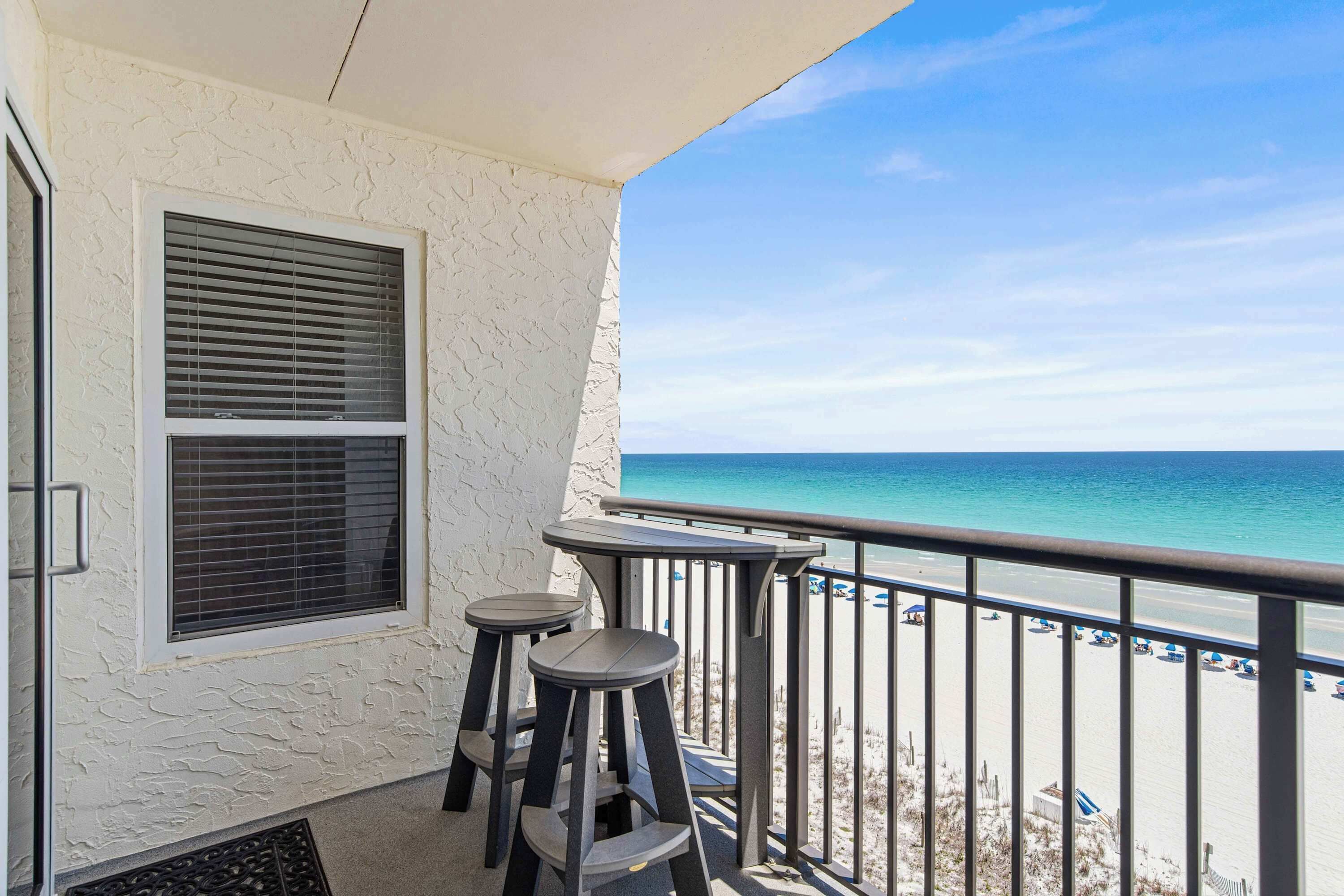 Un balcón con vista a la playa, una ventana con persianas y dos taburetes.