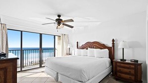 Quarto de hotel com uma cama grande, uma mesa de cabeceira de madeira, ventoinha no teto e vista para o oceano através de uma porta de vidro deslizante.