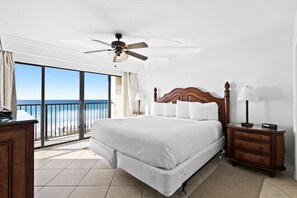 A hotel room with a large bed, a wooden nightstand, a ceiling fan, and a view of the ocean through a sliding glass door. - Large, updated condo - quick walk to beach & convenient to shopping (Miramar Beach)