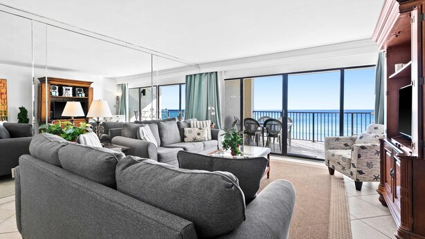 Sala de estar moderna com sofás cinzas, uma mesa de centro e uma TV. Há uma varanda com vista para o mar.
